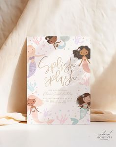 a card with mermaids on it sitting next to a white sheet and pillow in the background