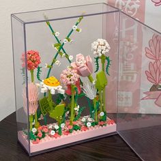 a glass case with flowers and plants inside it on a table next to a pink wall