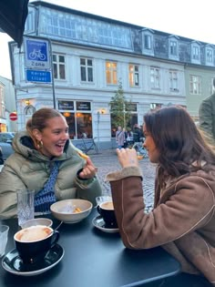 two women sitting at an outdoor table eating food and talking to each other, with buildings in the background