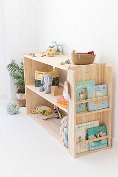 a wooden book shelf with books and toys on it in a white room next to a potted plant