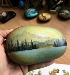 a hand holding a painted rock with trees and mountains in the background on top of a wooden table