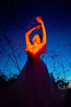 a woman in a long dress holding her arms up with the sun setting behind her