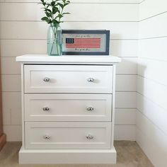 a white dresser with an american flag on top and flowers in a glass vase next to it