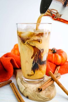 an iced drink being poured into a glass with cinnamon sticks and pumpkins in the background