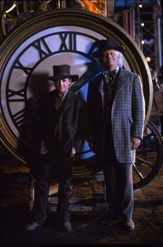 two men standing next to each other in front of a large clock with roman numerals