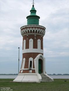 a large brick tower with a green top on the grass near water and clouds in the sky