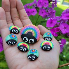 a person is holding some colorful rocks in their hand and purple flowers behind them with eyes painted on them