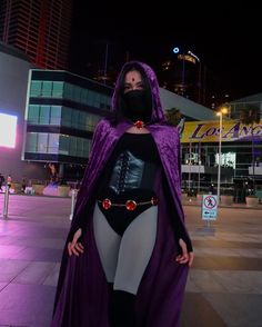 a woman in a purple cape and mask standing on the street at night with her hands on her hips