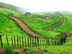 a grassy hill with houses on it and a wooden fence in the foreground, surrounded by green hills