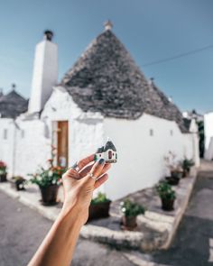 a hand holding a tiny toy house in front of a white building with potted plants