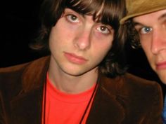 two young men standing next to each other in front of a dark background, one wearing a hat and the other looking at the camera