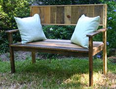 a wooden bench with two pillows sitting on it's back and in front of some bushes