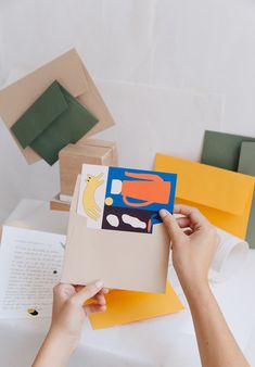 someone is holding up an open book on a table with many different colored papers and envelopes