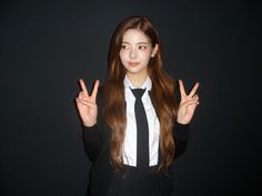 a woman in a tie and dress shirt making the peace sign with her hands while standing against a black background