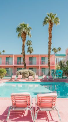 two lawn chairs sitting in front of a swimming pool with palm trees and pink buildings