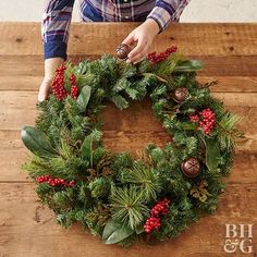 a woman is decorating a christmas wreath on a wooden table with greenery and pine cones