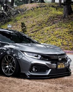 the front end of a gray car parked on top of a dirt road next to trees