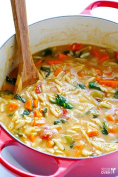 a red pot filled with pasta and vegetables next to a wooden spoon on top of a table