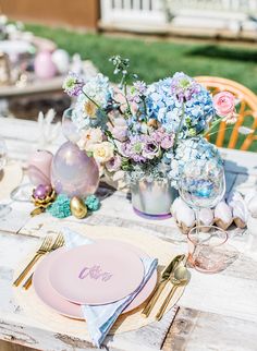 an outdoor table set with pink and blue plates, silverware and flowers in vases