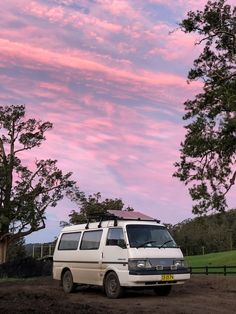 a white van parked on top of a dirt field under a pink sky with clouds