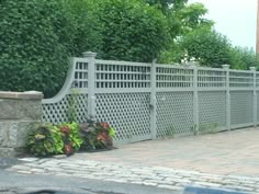 a white fence with flowers growing on the side of it and trees in the background