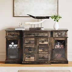 an old wooden sideboard with glass doors and drawers in front of a painting on the wall