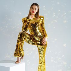 a woman in gold sequins poses on a white pedestal