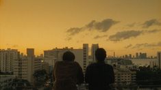 two people sitting on top of a roof watching the sun go down over a city