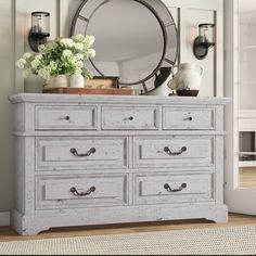 a white dresser with flowers on top and a round mirror above it in a room