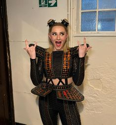 a woman in black and orange outfit standing next to a white wall with her hands up