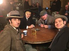 three men sitting at a table with beer in front of them and people standing around