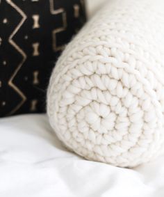 a white knitted pillow sitting on top of a bed next to a black pillow