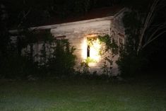 a small white house sitting in the middle of a forest at night with light coming from it's window