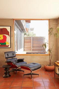 an eames chair and ottoman sit in front of a window with a painting on the wall