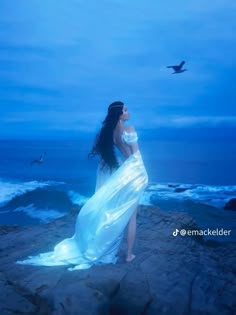 a woman in a white dress standing on rocks near the ocean with a bird flying overhead