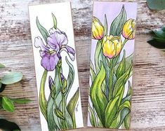 two bookmarks with flowers painted on them sitting next to some green leaves and plants