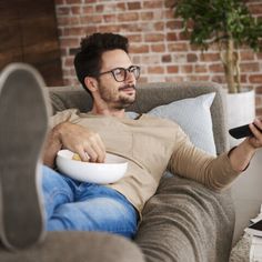 a man sitting on a couch holding a remote control and bowl of popcorn in his hand