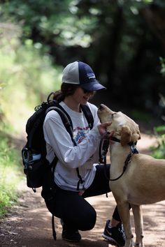 a woman kneeling down next to a brown dog on a dirt road with trees in the background