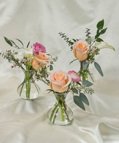 three vases filled with different types of flowers on a white cloth covered tablecloth