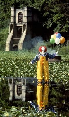 a clown is standing in the water with some balloons on his head and wearing yellow overalls