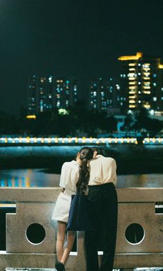 two people are sitting on a bench looking at the water and buildings in the background