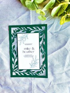 a green and white wedding card sitting on top of a blue blanket next to a plant