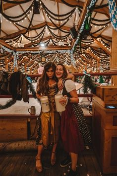 two women standing next to each other in front of a wooden structure with lights on it