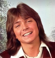 a young man with long hair smiling at the camera while wearing a red vest and white shirt