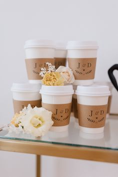 six coffee cups sitting on top of a glass table with flowers in the middle and monogrammed labels