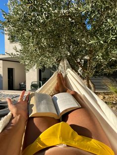 a person laying in a hammock reading a book under an olive tree with their feet propped up