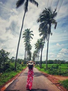 a woman in a long dress and straw hat walking down the road with palm trees behind her