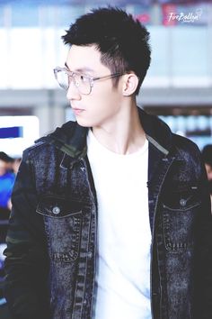 a young man wearing glasses and a denim jacket looks off into the distance while standing in an airport