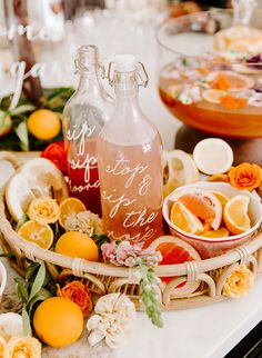 an arrangement of fruit and wine bottles on a table with flowers, oranges and lemons