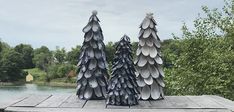 three metal trees sitting on top of a wooden table next to some water and trees